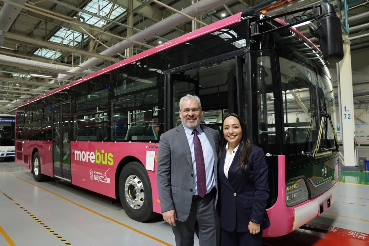 Dos personas de pie y sonriendo frente a un autobús rosa dentro de una nave industrial.