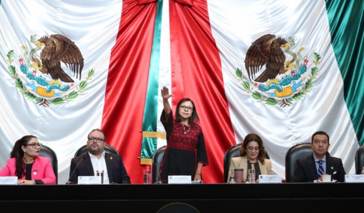 Cinco personas están sentadas en una mesa frente a una gran bandera de México, mientras una mujer en el centro levanta la mano derecha.
