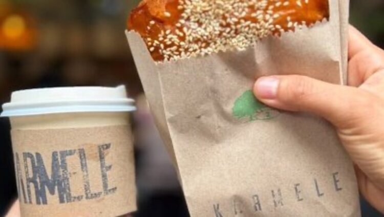 Una mano sostiene una bebida caliente y otra un pan cubierto de ajonjolí en una bolsa de papel.