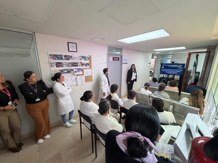 Un grupo de personas, algunas con batas blancas, asiste a una presentación en una sala pequeña con sillas y un monitor.
