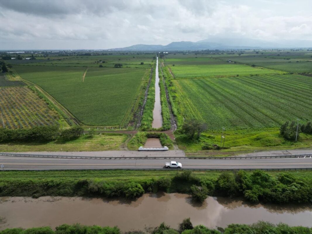 Un paisaje rural con campos verdes, un canal de agua recto, una carretera con un auto blanco y un río en primer plano bajo un cielo nublado.