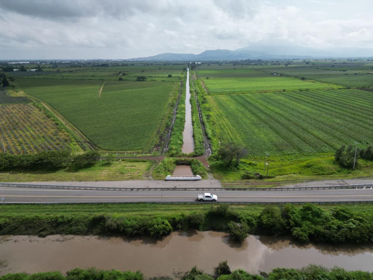 Un paisaje rural con campos verdes, un canal de agua recto, una carretera con un auto blanco y un río en primer plano bajo un cielo nublado.