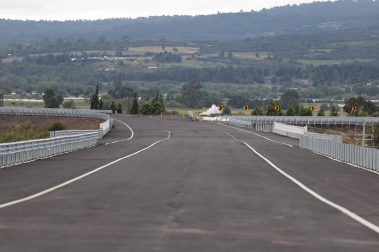 Una carretera vacía con barandales metálicos rodeada de colinas y vegetación al fondo.