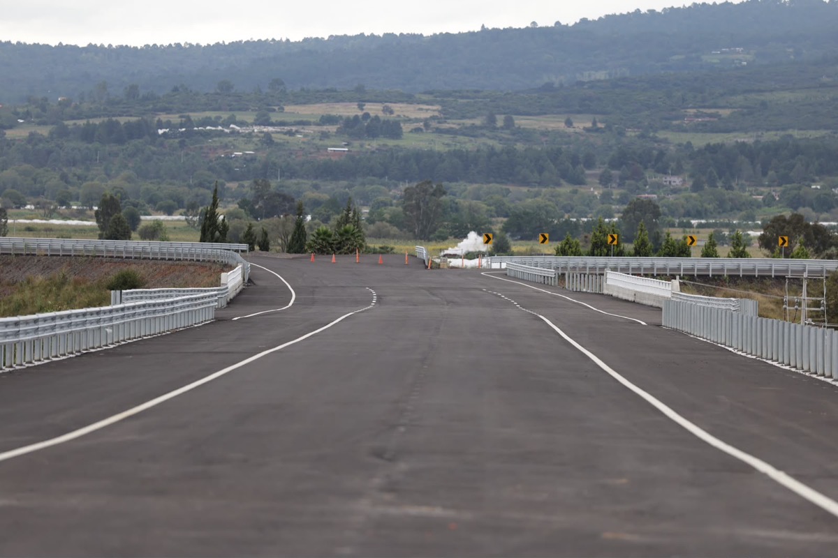 Una carretera vacía con barandales metálicos rodeada de colinas y vegetación al fondo.