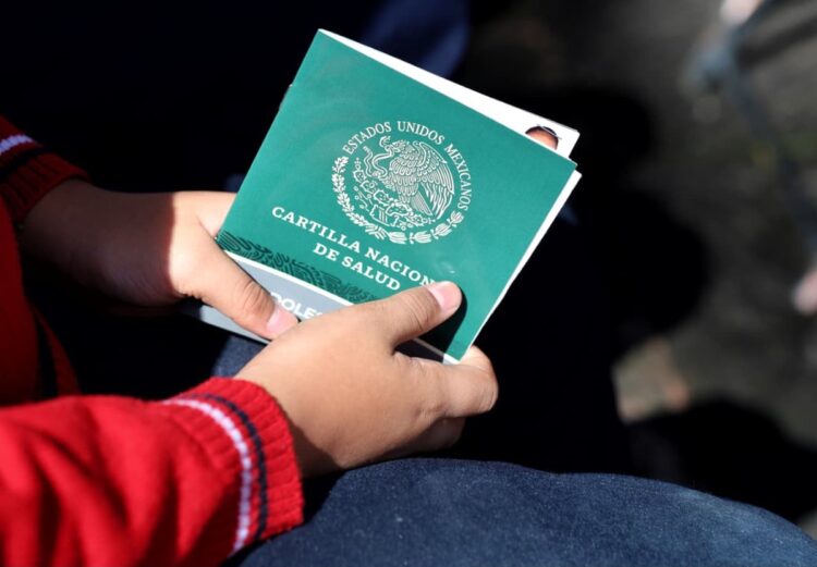 Unas manos sostienen una cartilla verde de salud mexicana con el escudo nacional en la portada.