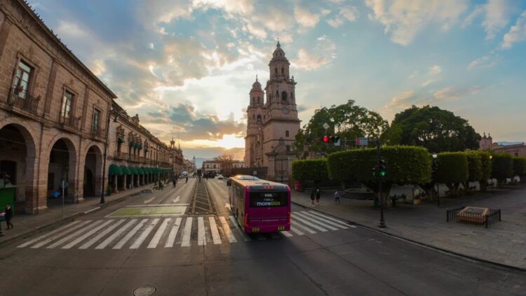 Un autobús circula por una calle amplia con edificios coloniales y una catedral al fondo al atardecer.