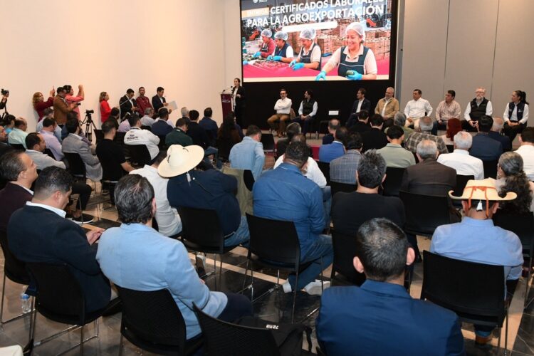 Un grupo de personas asiste a una conferencia en un auditorio, con varios panelistas sentados en el escenario y una pantalla grande mostrando trabajadores en una línea de producción.