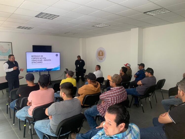 Un grupo de personas está sentado en una sala mientras un hombre da una presentación con una pantalla que muestra información sobre certificación vehicular.
