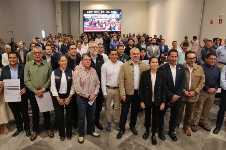 Un grupo de personas posa para una foto en un salón durante un evento, con una pantalla al fondo y varias filas de asistentes sentados y de pie.