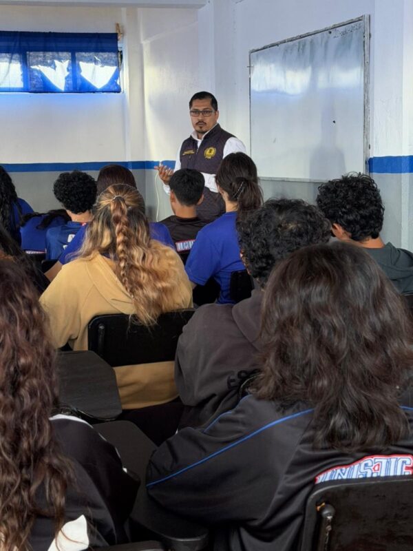 Un hombre está dando una clase frente a un grupo de estudiantes sentados en un salón con un pizarrón blanco.