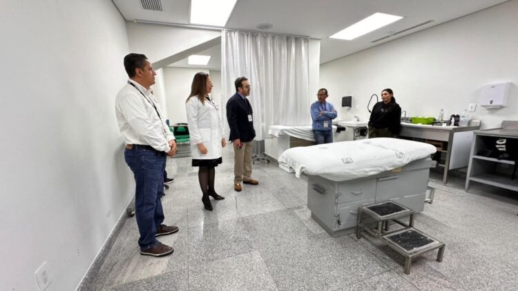 Varias personas están de pie en una sala blanca con una camilla médica y equipo clínico.