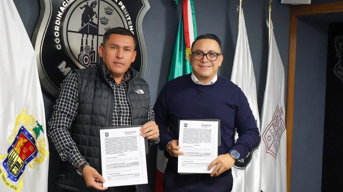Dos hombres posan sonrientes sosteniendo documentos frente a varias banderas y un emblema institucional en la pared.
