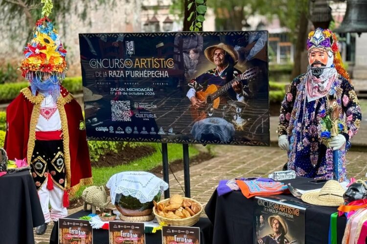 Dos personas con coloridos trajes tradicionales están junto a un cartel que anuncia un concurso artístico en Zacán, Michoacán, rodeadas de mesas con comida y objetos típicos.