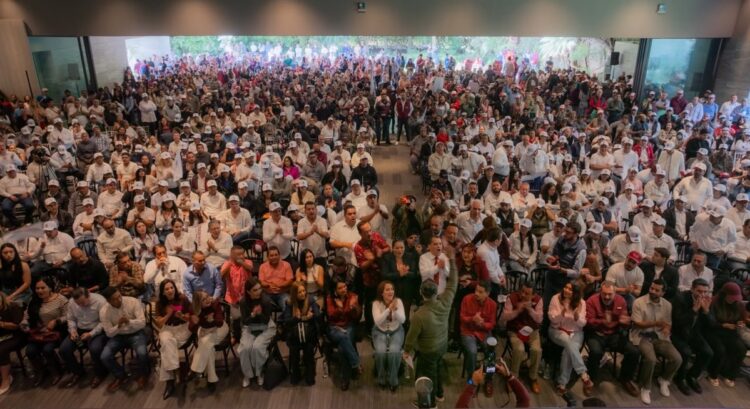 Una multitud de personas sentadas y de pie en un auditorio cerrado durante un evento.