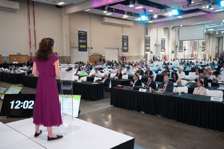 Una mujer de vestido morado habla en un podio frente a un público sentado en un salón de conferencias.