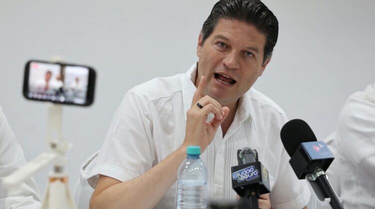 Un hombre con camisa blanca habla en una conferencia frente a micrófonos y una cámara de teléfono.