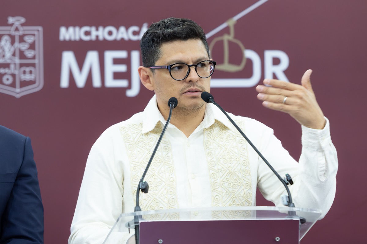 Hombre hablando en un micrófono con gafas, camiseta blanca bordada, gesticulando durante una conferencia en un podio. Fondo V.