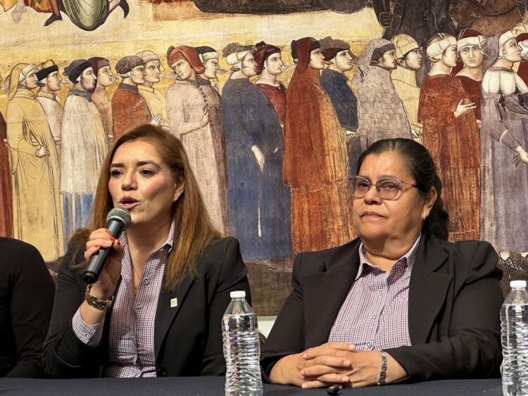 Dos mujeres con trajes formales están sentadas frente a un mural, una de ellas hablando con un micrófono y la otra con las manos cruzadas.