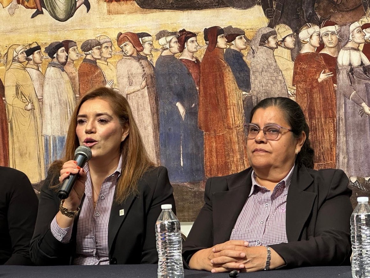 Mujeres políticas hablan en una mesa; una de ellas sostiene un micrófono, al fondo mural histórico.