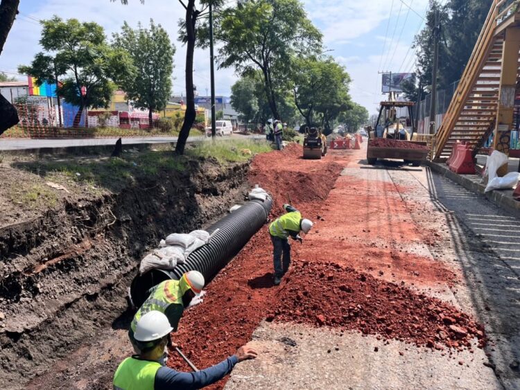 Trabajadores de construcción instalan una tubería grande y esparcen tierra roja en una calle en reparación.