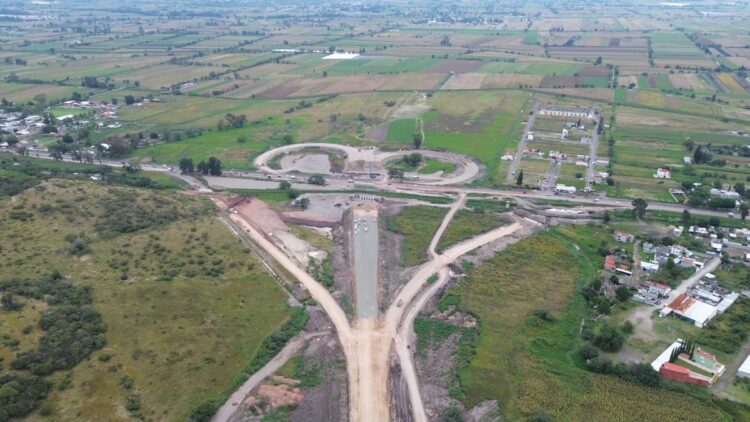 Vista aérea de una carretera en construcción que atraviesa campos y áreas rurales con algunas casas dispersas.