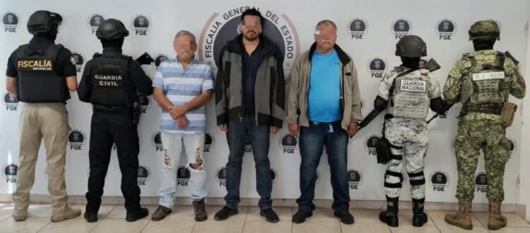 Tres hombres detenidos están flanqueados por oficiales armados de distintas corporaciones frente a un fondo de la Fiscalía General del Estado.