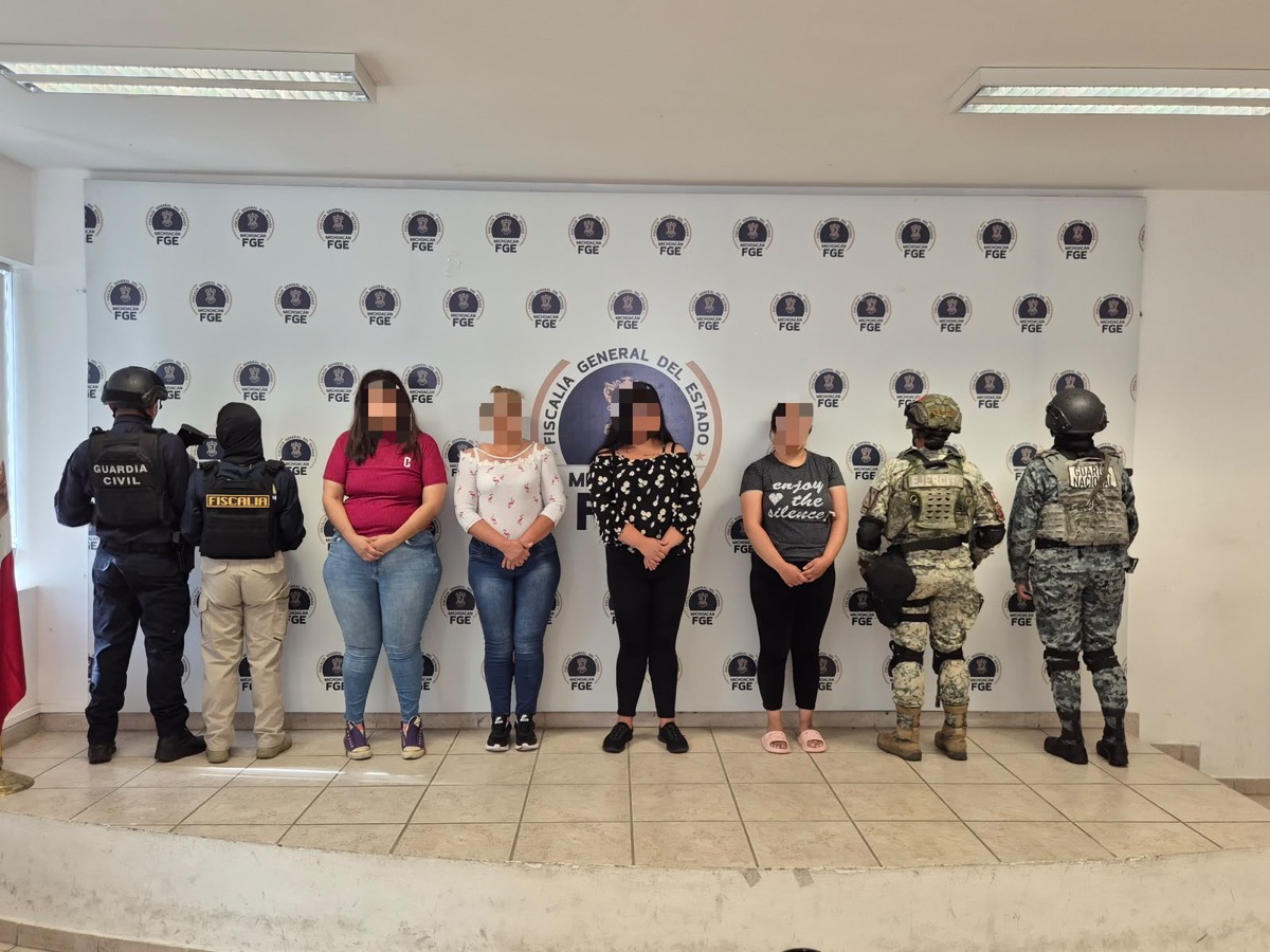 Cinco mujeres escoltadas por agentes armados posan frente a un fondo de la Fiscalía General del Estado.