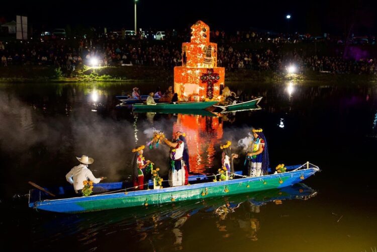 Varias personas en canoas decoradas navegan de noche frente a una estructura iluminada en un lago, con humo y público observando desde la orilla.