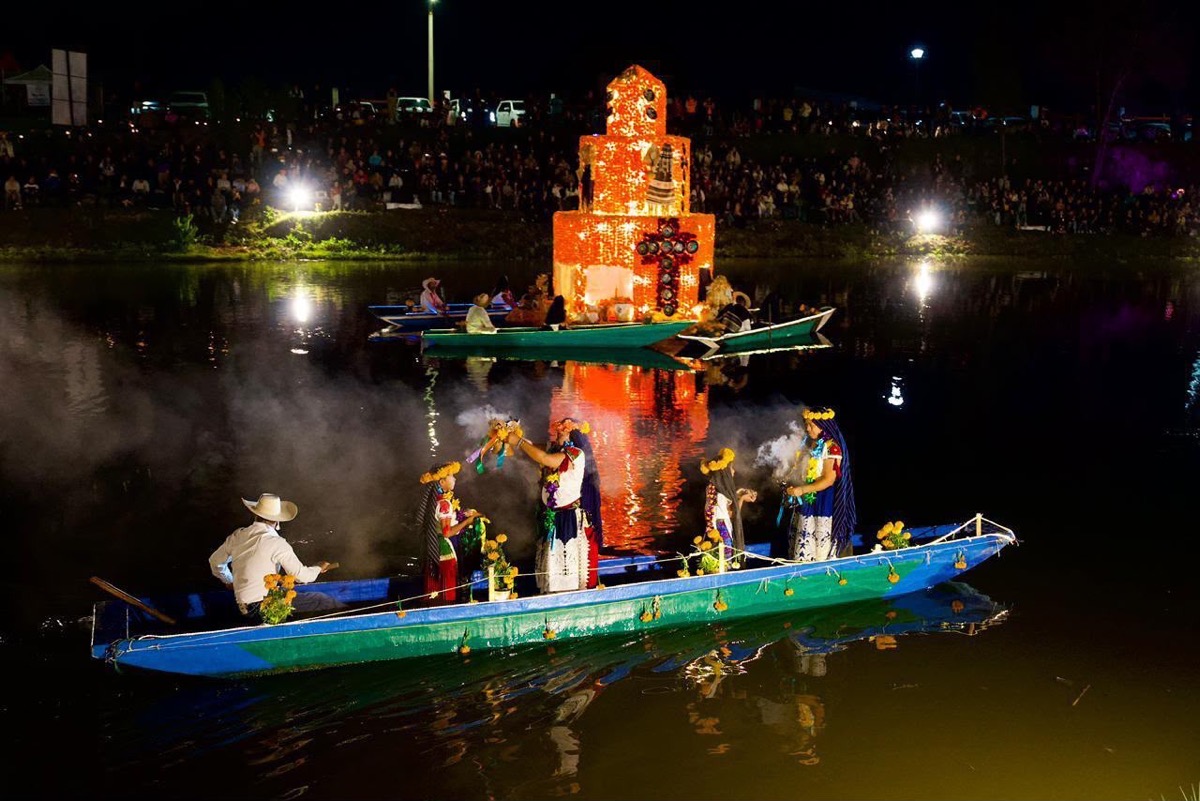 Varias personas en canoas decoradas navegan de noche frente a una estructura iluminada en un lago, con humo y público observando desde la orilla.