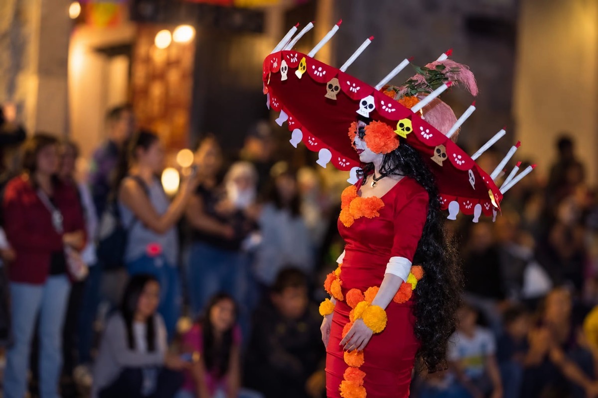 Figura en traje rojo con sombrero decorado y flores naranjas durante desfile público; público borroso al fondo en una calle.