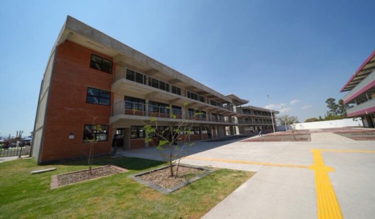 Edificio escolar de tres pisos con áreas verdes y un amplio patio de concreto bajo un cielo despejado.