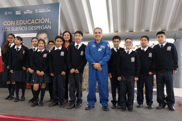 Un grupo de estudiantes con uniforme escolar posa junto a un hombre con traje espacial azul en un escenario interior.
