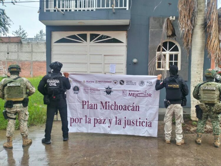 Cuatro agentes armados están de espaldas sosteniendo una lona frente a una casa que dice “Plan Michoacán por la paz y la justicia”.