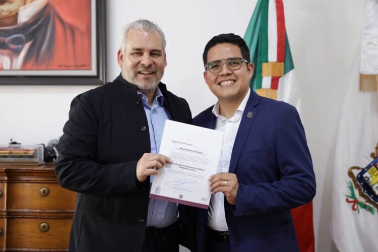 Dos hombres sonrientes posan juntos mientras sostienen un documento frente a una bandera y un mueble de madera.