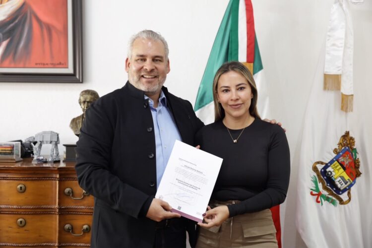 Dos personas sonrientes posan juntas en una oficina mientras sostienen un documento frente a banderas y un escritorio.