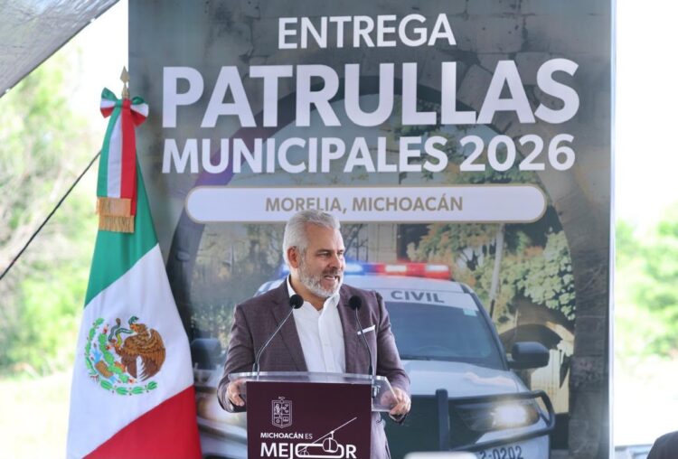 Un hombre de cabello canoso habla en un podio frente a un cartel que dice “Entrega Patrullas Municipales 2026” en Morelia, Michoacán, con una patrulla y una bandera de México a su lado.