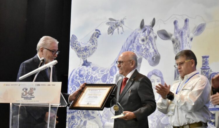 Tres hombres están en un escenario durante una ceremonia, donde uno de ellos recibe un diploma y un trofeo mientras otro aplaude.