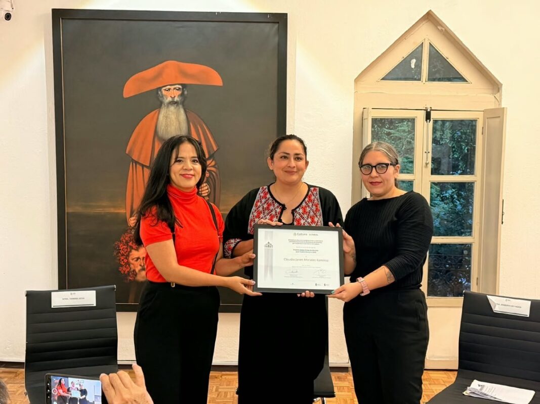Tres mujeres posan en una sala mientras sostienen un diploma, con una pintura y una ventana al fondo.