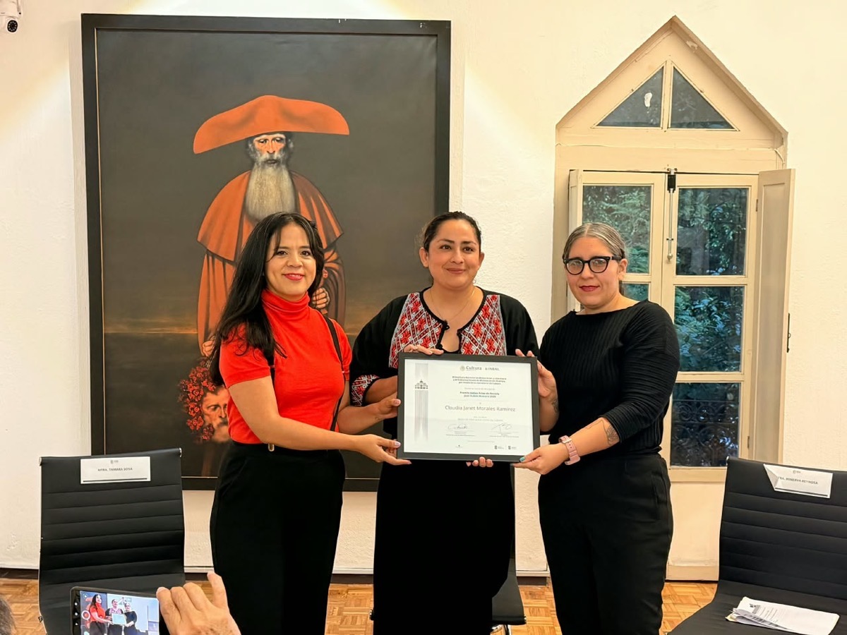 Tres mujeres posan en una sala mientras sostienen un diploma, con una pintura y una ventana al fondo.