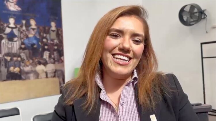 Mujer sonriente con saco negro y camisa a cuadros posando en una sala con un cuadro y un ventilador en la pared.