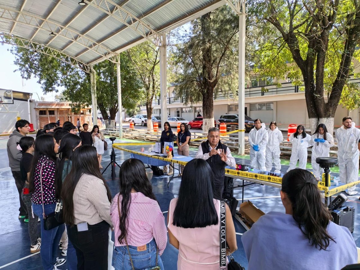 Un grupo de personas observa a un hombre hablando en una cancha techada, mientras otros llevan trajes blancos protectores detrás de una cinta amarilla.