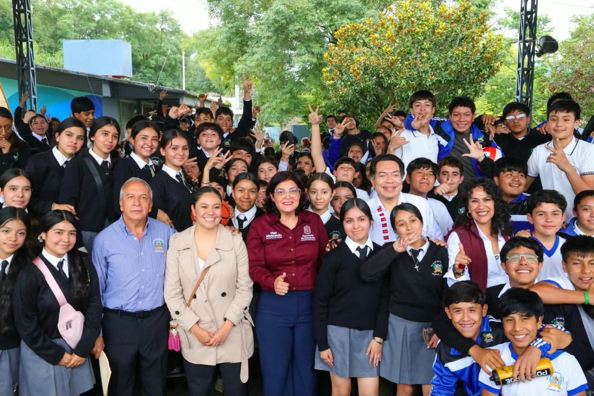 Un grupo grande de estudiantes y adultos posa sonriente al aire libre, con árboles y edificios escolares de fondo.