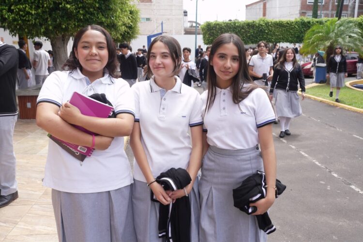 Tres estudiantes con uniforme escolar posan al aire libre mientras otros alumnos caminan en el fondo.