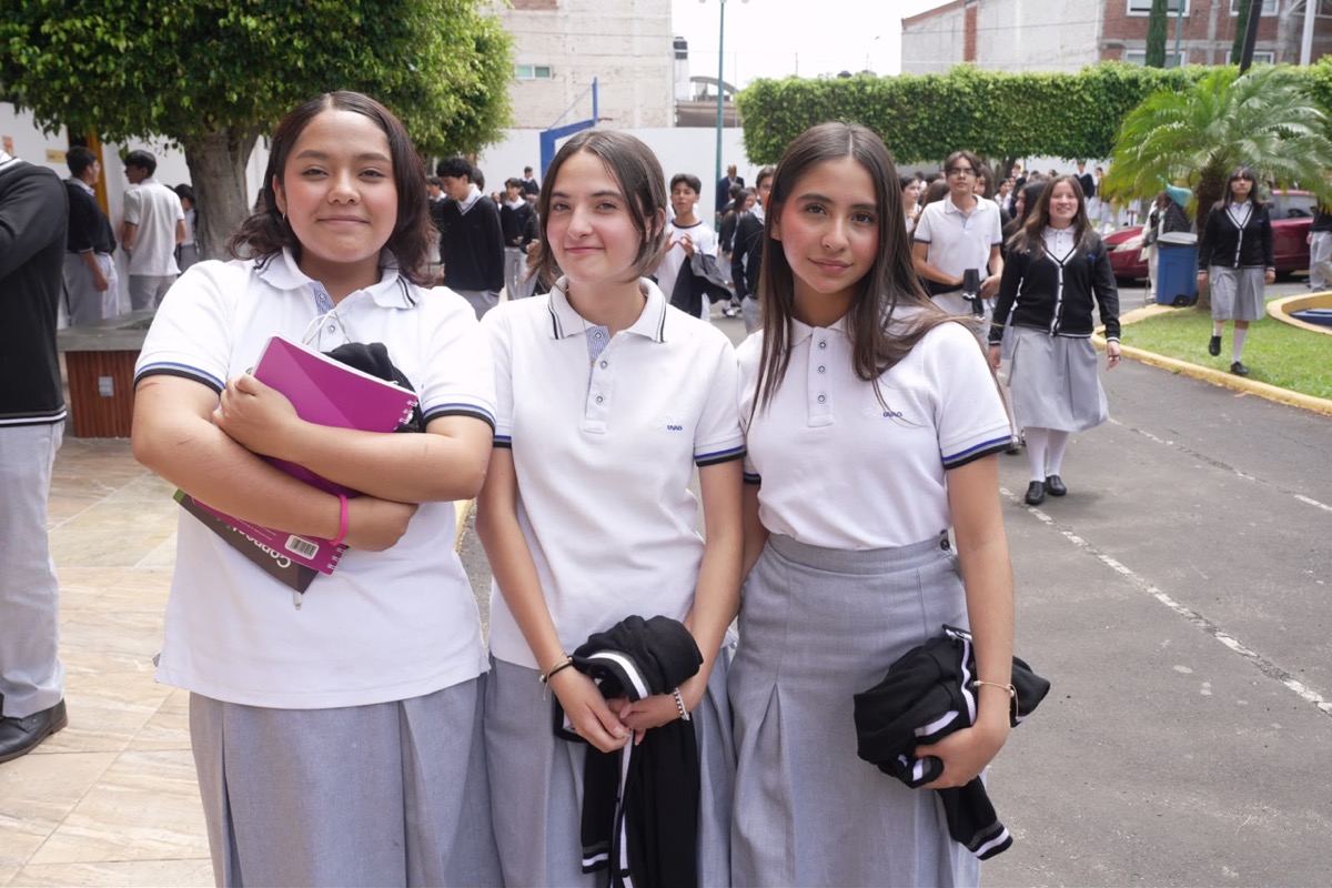 Tres estudiantes con uniforme escolar posan al aire libre mientras otros alumnos caminan en el fondo.