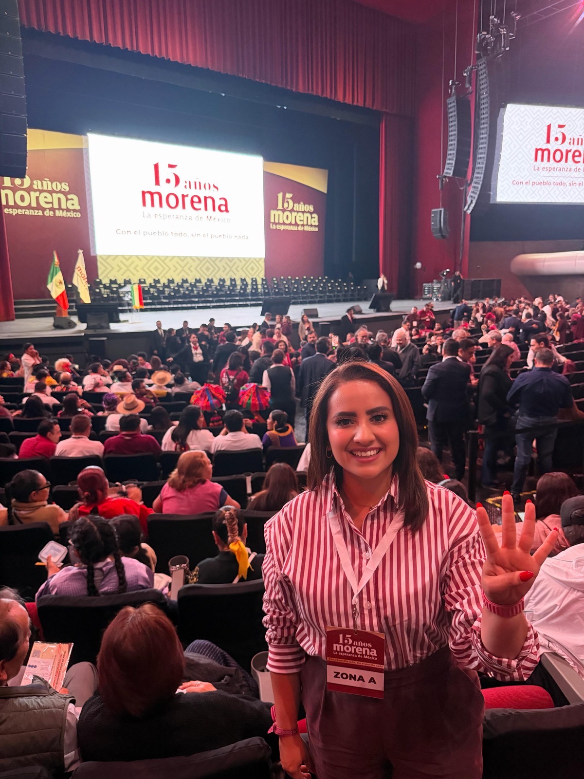 Una mujer sonriente con gafete de “Zona A” posa en un auditorio lleno de gente frente a un escenario con pantallas que dicen “15 años Morena”.