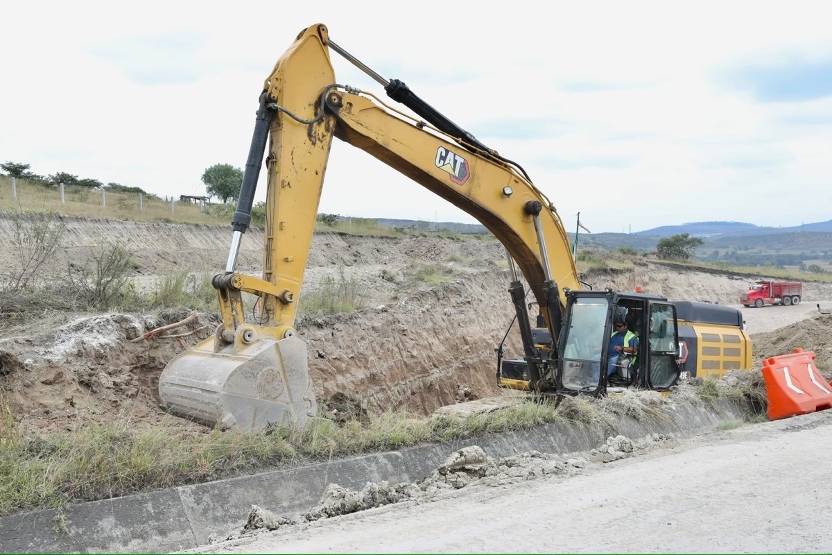 Una excavadora amarilla está cavando una zanja en una obra vial mientras un trabajador la opera.