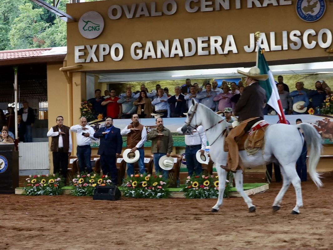 Un hombre a caballo porta una bandera mexicana frente a un grupo de personas en un evento en un recinto cubierto con flores y letreros.