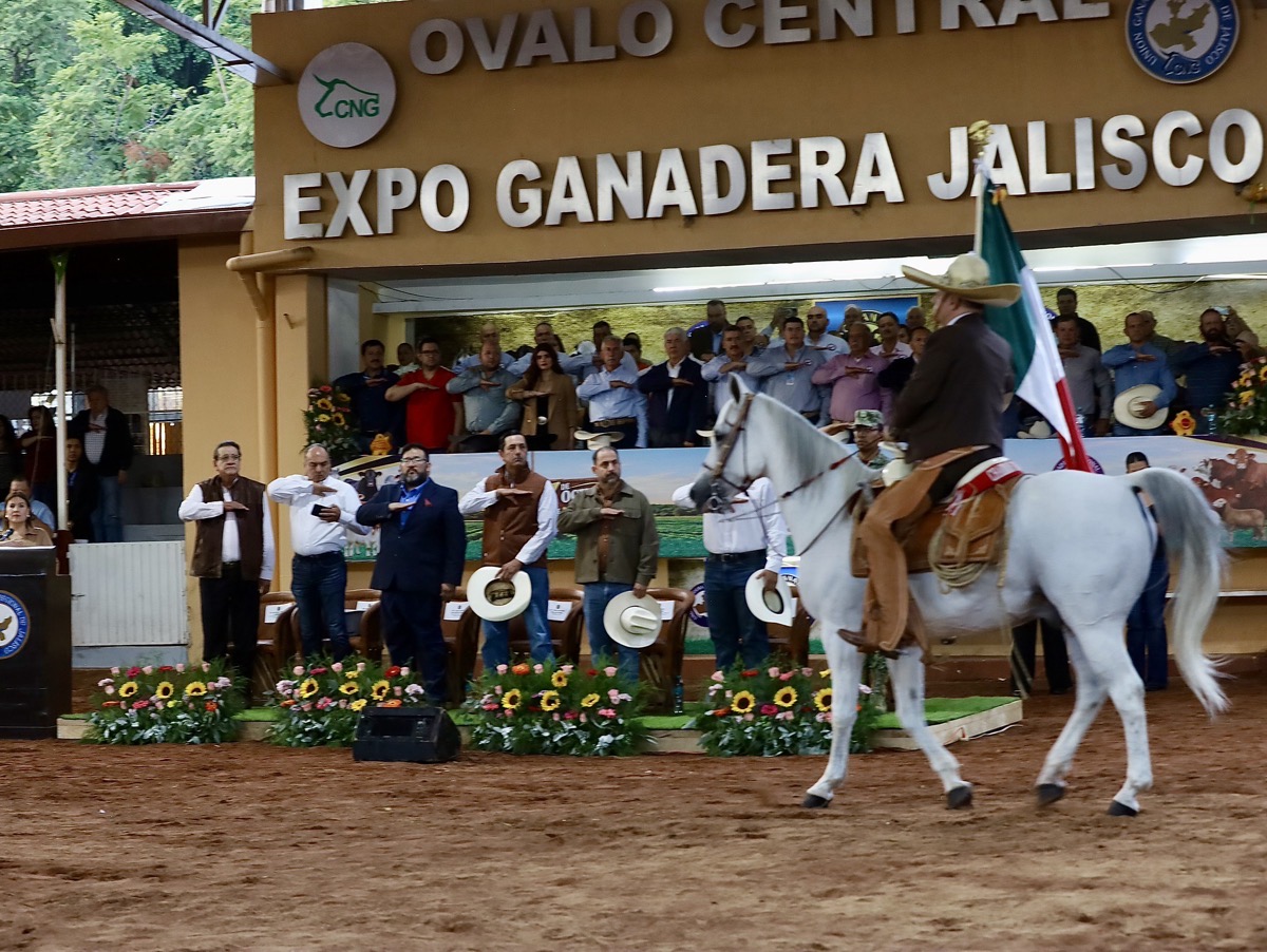 Un hombre a caballo porta una bandera mexicana frente a un grupo de personas en un evento en un recinto cubierto con flores y letreros.