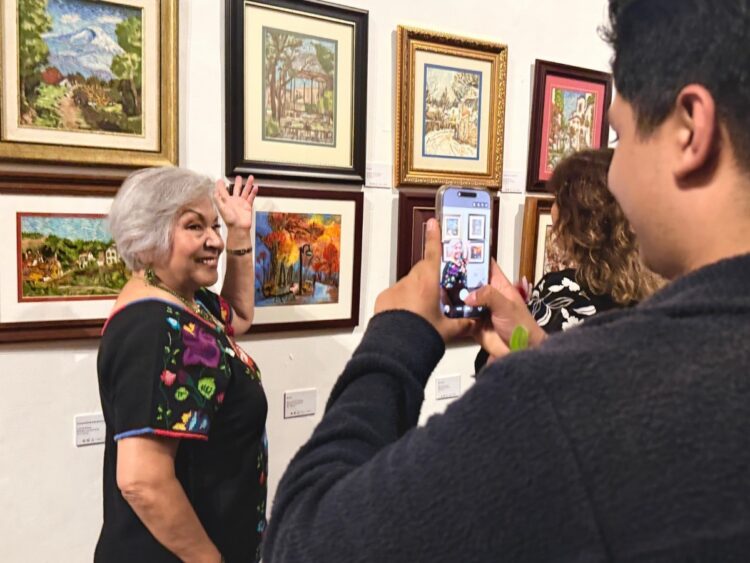 Una mujer sonríe y posa junto a varias pinturas en una galería mientras otra persona le toma una foto con su teléfono.