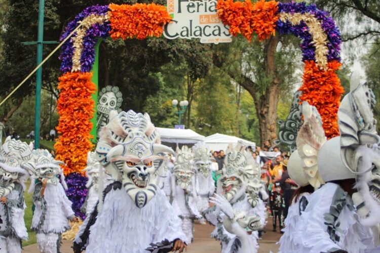 Desfile de máscaras blancas con trajes blancos y carcasas decorativas en un arco floral naranja y morado, durante una feria.